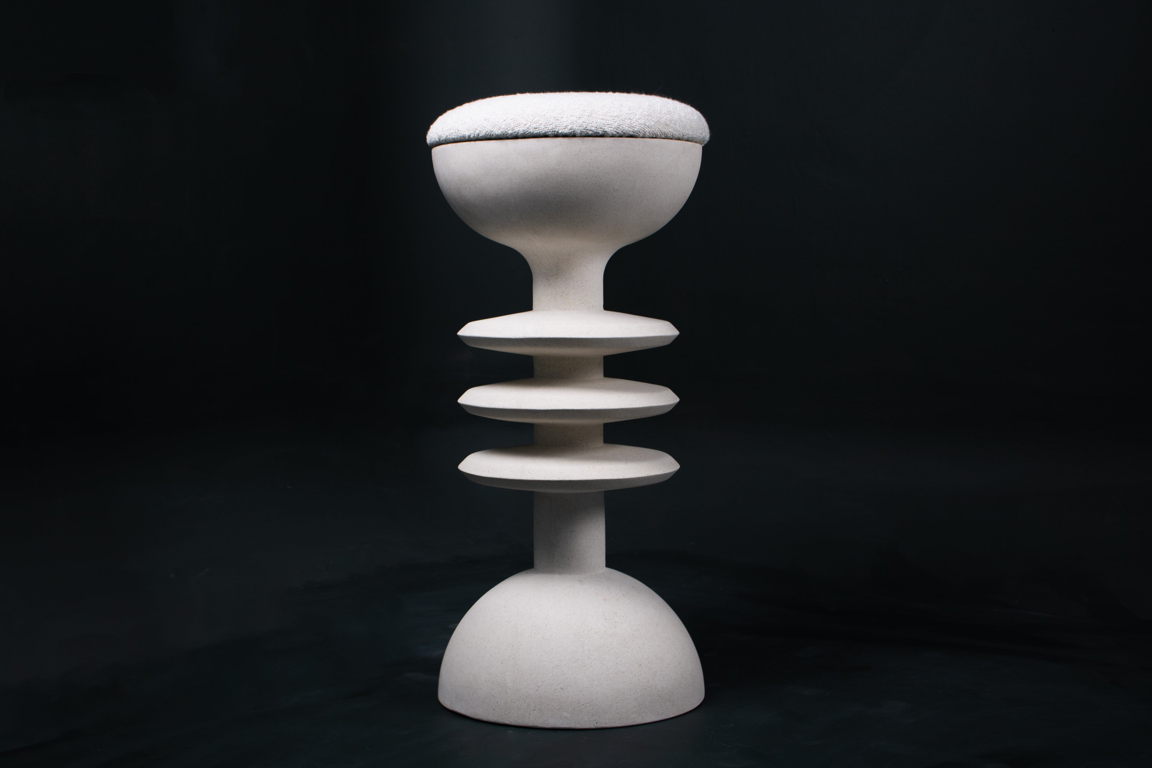 The Annulet barstool in white. The sculptural base rises from a domed foot through three stacked horizontal discs, narrowing to a stem that supports a wide chalice-shaped seat cradle, topped with a round white bouclé upholstered seat.