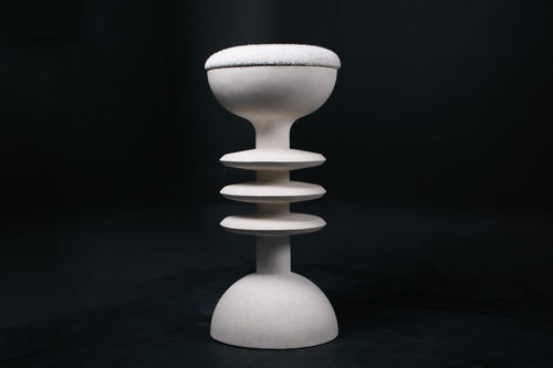 The Annulet barstool in white. The sculptural base rises from a domed foot through three stacked horizontal discs, narrowing to a stem that supports a wide chalice-shaped seat cradle, topped with a round white bouclé upholstered seat.