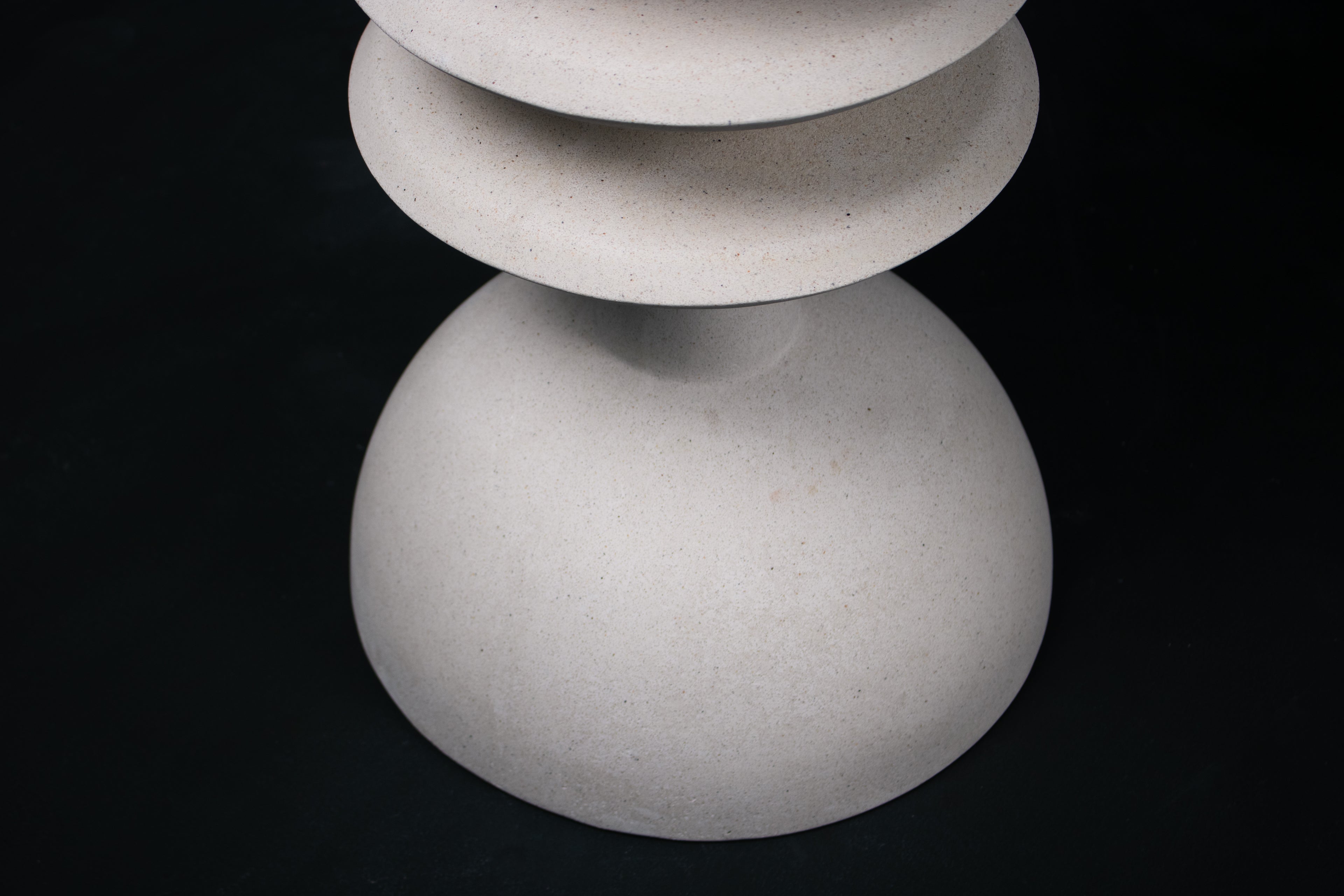 The Annulet barstool in white. The sculptural base rises from a domed foot through three stacked horizontal discs, narrowing to a stem that supports a wide chalice-shaped seat cradle, topped with a round white bouclé upholstered seat.