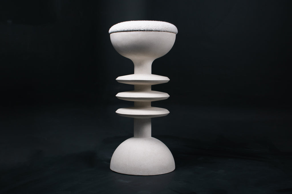 The Annulet barstool in white. The sculptural base rises from a domed foot through three stacked horizontal discs, narrowing to a stem that supports a wide chalice-shaped seat cradle, topped with a round white bouclé upholstered seat.