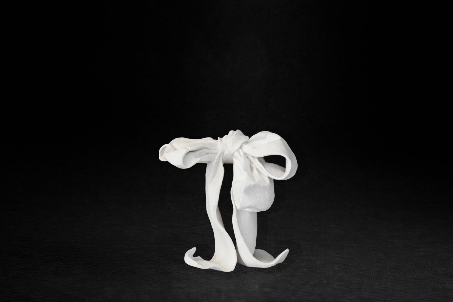 A white plaster sculpture depicting a loosely tied bow. The ribbon forms billow and curl freely, capturing the movement and drape of fabric frozen mid-gesture, with trailing ends pooling at the base.