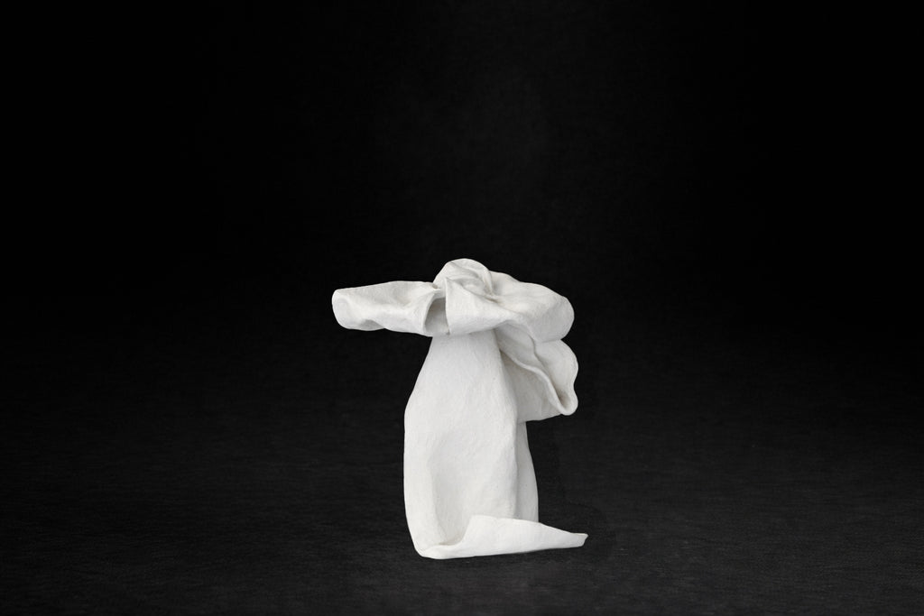 A white plaster sculpture depicting a loosely tied bow. The ribbon forms billow and curl freely, capturing the movement and drape of fabric frozen mid-gesture, with trailing ends pooling at the base.