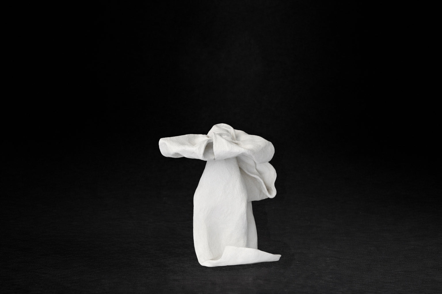 A white plaster sculpture depicting a loosely tied bow. The ribbon forms billow and curl freely, capturing the movement and drape of fabric frozen mid-gesture, with trailing ends pooling at the base.