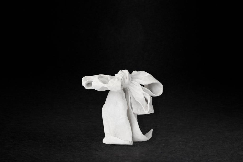 A white plaster sculpture depicting a loosely tied bow. The ribbon forms billow and curl freely, capturing the movement and drape of fabric frozen mid-gesture, with trailing ends pooling at the base.
