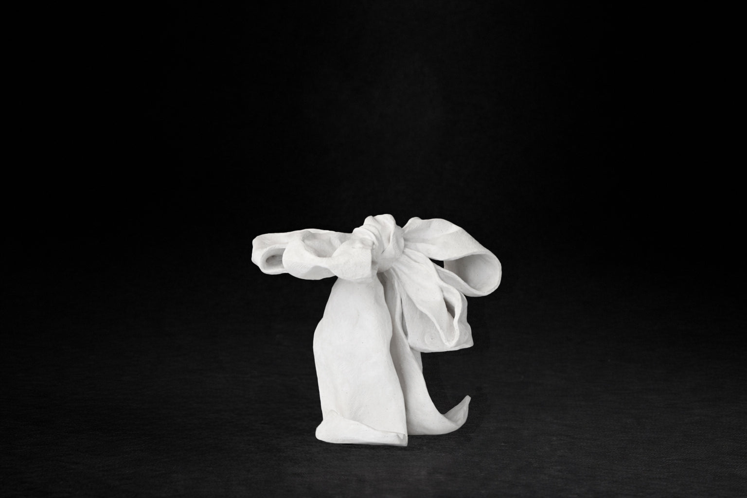 A white plaster sculpture depicting a loosely tied bow. The ribbon forms billow and curl freely, capturing the movement and drape of fabric frozen mid-gesture, with trailing ends pooling at the base.
