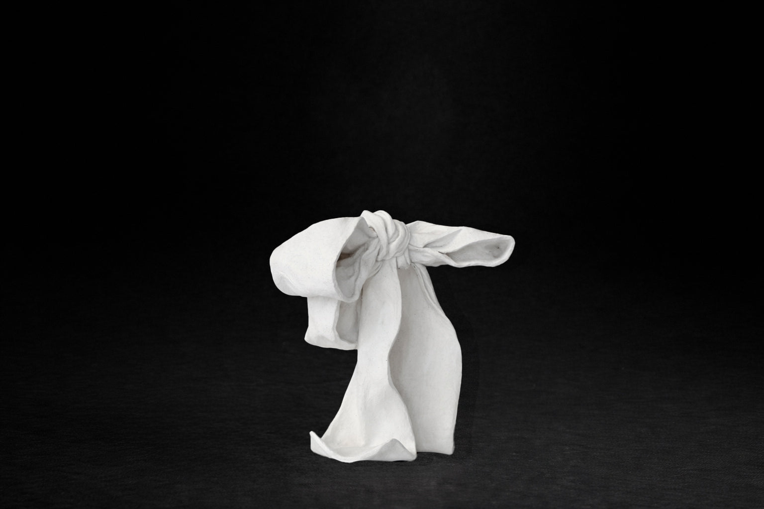 A white plaster sculpture depicting a loosely tied bow. The ribbon forms billow and curl freely, capturing the movement and drape of fabric frozen mid-gesture, with trailing ends pooling at the base.