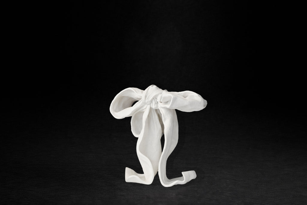 A white plaster sculpture depicting a loosely tied bow. The ribbon forms billow and curl freely, capturing the movement and drape of fabric frozen mid-gesture, with trailing ends pooling at the base.