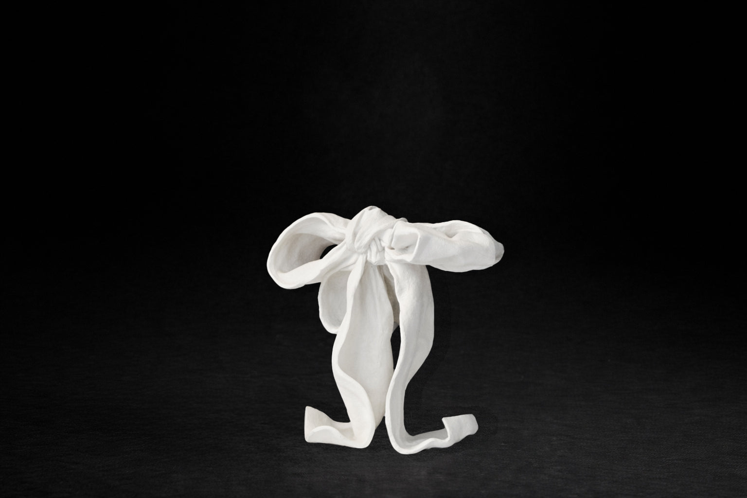 A white plaster sculpture depicting a loosely tied bow. The ribbon forms billow and curl freely, capturing the movement and drape of fabric frozen mid-gesture, with trailing ends pooling at the base.