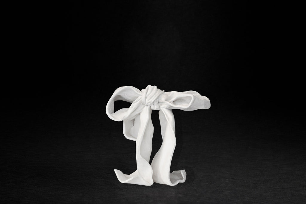 A white plaster sculpture depicting a loosely tied bow. The ribbon forms billow and curl freely, capturing the movement and drape of fabric frozen mid-gesture, with trailing ends pooling at the base.