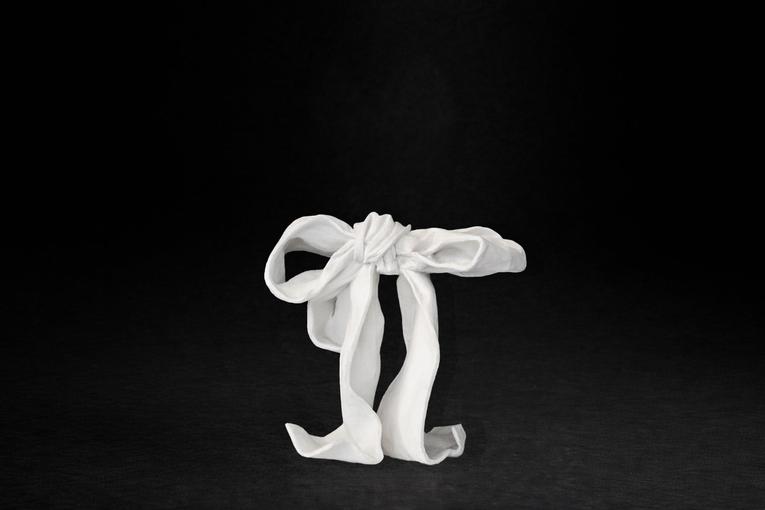 A white plaster sculpture depicting a loosely tied bow. The ribbon forms billow and curl freely, capturing the movement and drape of fabric frozen mid-gesture, with trailing ends pooling at the base.