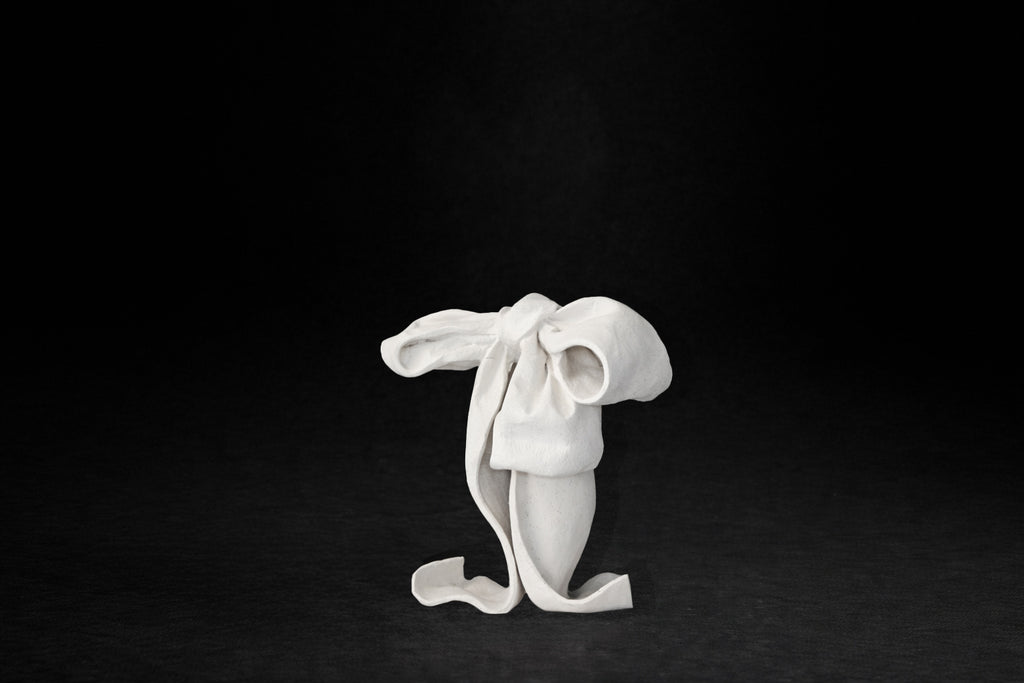 A white plaster sculpture depicting a loosely tied bow. The ribbon forms billow and curl freely, capturing the movement and drape of fabric frozen mid-gesture, with trailing ends pooling at the base.