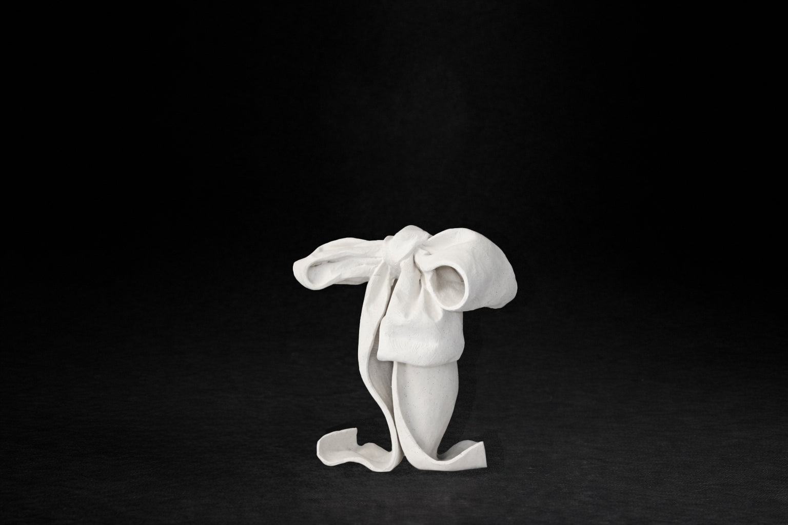 A white plaster sculpture depicting a loosely tied bow. The ribbon forms billow and curl freely, capturing the movement and drape of fabric frozen mid-gesture, with trailing ends pooling at the base.