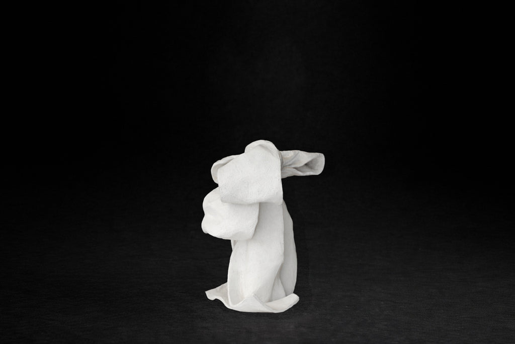 A white plaster sculpture depicting a loosely tied bow. The ribbon forms billow and curl freely, capturing the movement and drape of fabric frozen mid-gesture, with trailing ends pooling at the base.