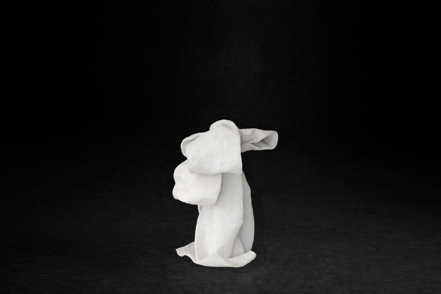 A white plaster sculpture depicting a loosely tied bow. The ribbon forms billow and curl freely, capturing the movement and drape of fabric frozen mid-gesture, with trailing ends pooling at the base.