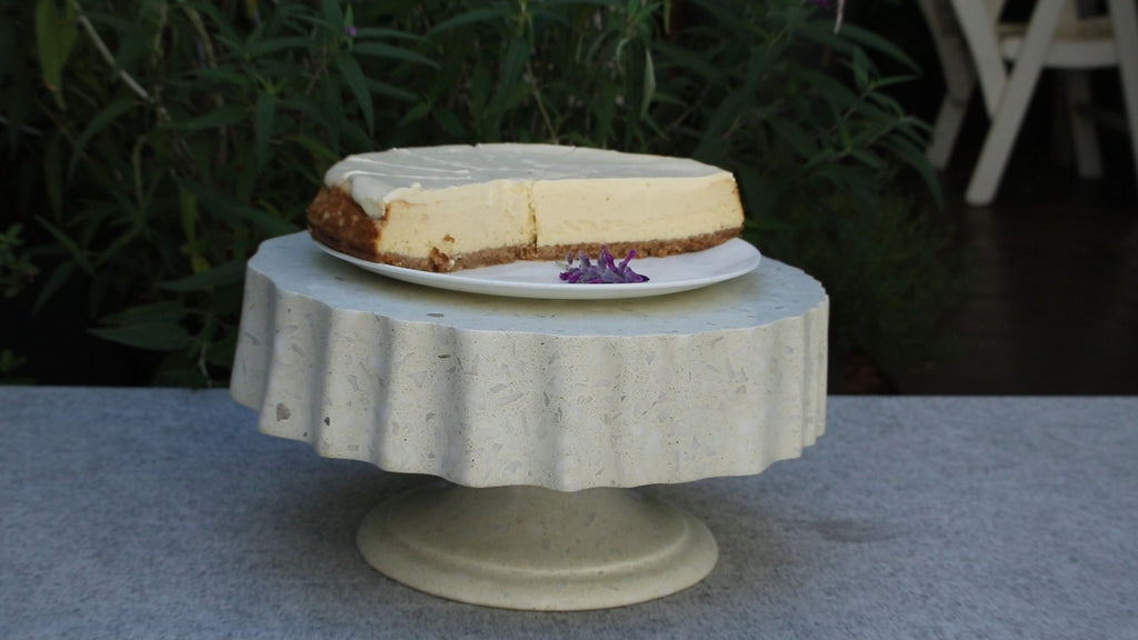 The Cascade cake stand in terrazzo. The substantial circular platform has a deeply fluted edge with vertical ribbing running around the full circumference, resting on a smooth tapered pedestal foot. The pale grey terrazzo surface is flecked with fragments of white, blush, and terracotta quartz.