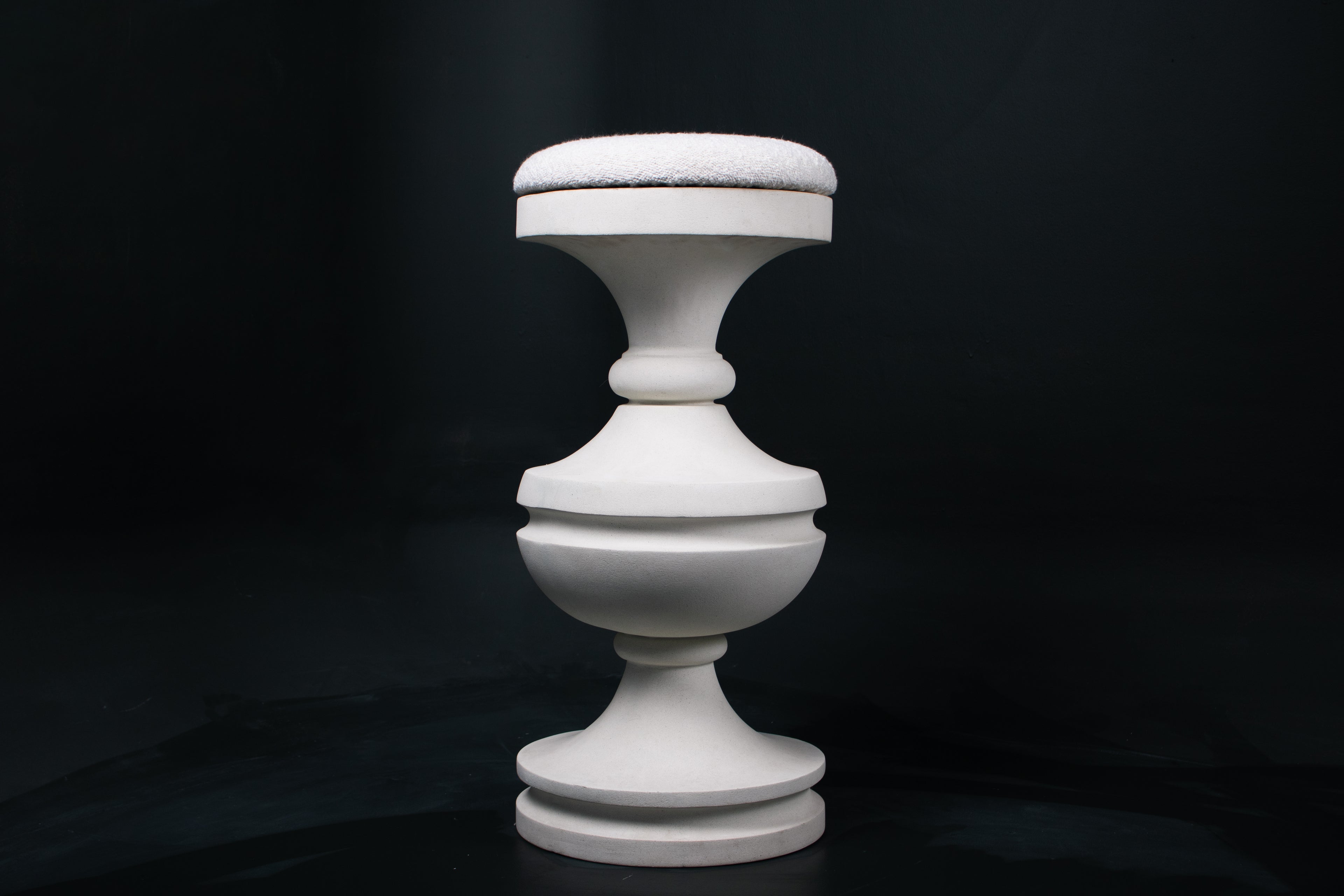 Chalice barstool in white. The substantial turned base draws on classical column and urn forms, a layered circular foot rises through a bulbous lower body, pinches to a narrow waist, then flares outward to support a round white bouclé upholstered seat.