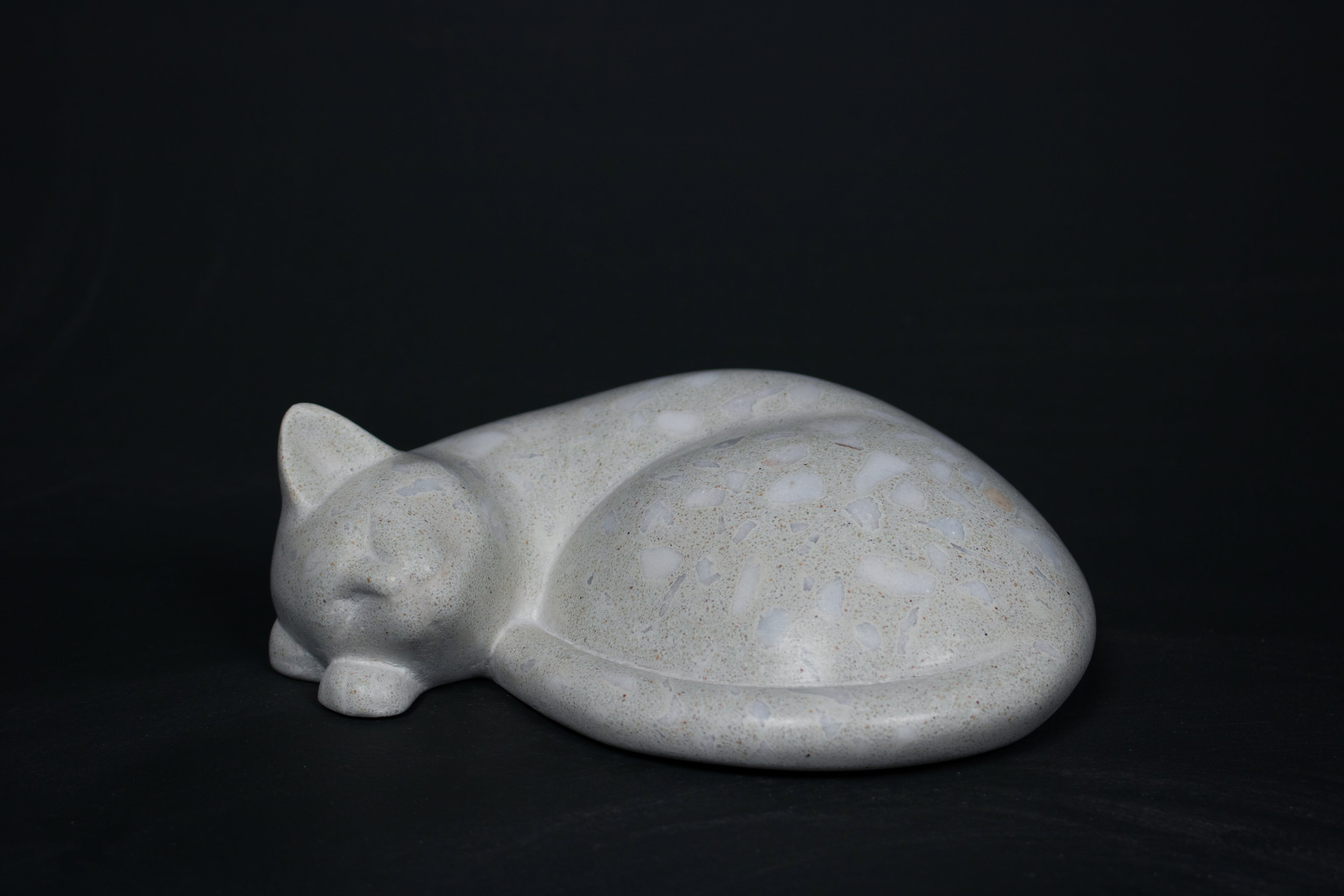 The Curly Cat terrazzo doorstop. A close-up view reveals the smoothly cast sleeping cat form, head resting on tucked paws, eyes closed, body curled into a low rounded mass. The pale grey terrazzo surface is flecked with irregular fragments of white and blush quartz.