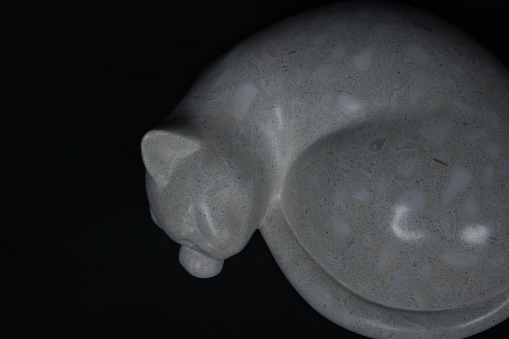 The Curly Cat terrazzo doorstop. A close-up view reveals the smoothly cast sleeping cat form, head resting on tucked paws, eyes closed, body curled into a low rounded mass. The pale grey terrazzo surface is flecked with irregular fragments of white and blush quartz.