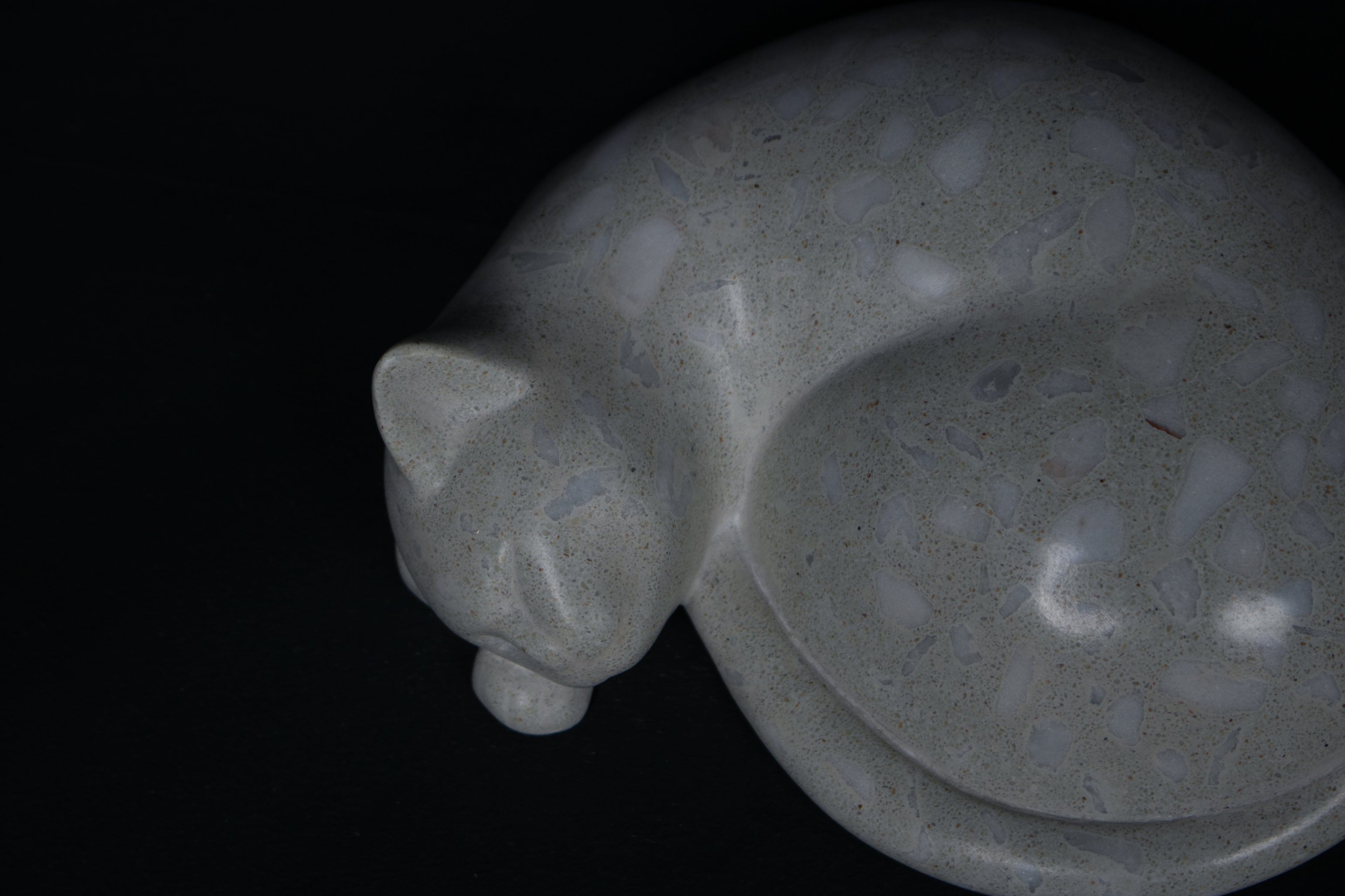 The Curly Cat terrazzo doorstop. A close-up view reveals the smoothly cast sleeping cat form, head resting on tucked paws, eyes closed, body curled into a low rounded mass. The pale grey terrazzo surface is flecked with irregular fragments of white and blush quartz.