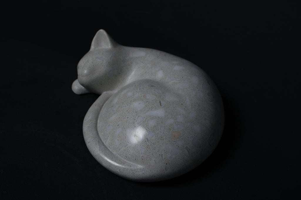 The Curly Cat terrazzo doorstop. A close-up view reveals the smoothly cast sleeping cat form, head resting on tucked paws, eyes closed, body curled into a low rounded mass. The pale grey terrazzo surface is flecked with irregular fragments of white and blush quartz.