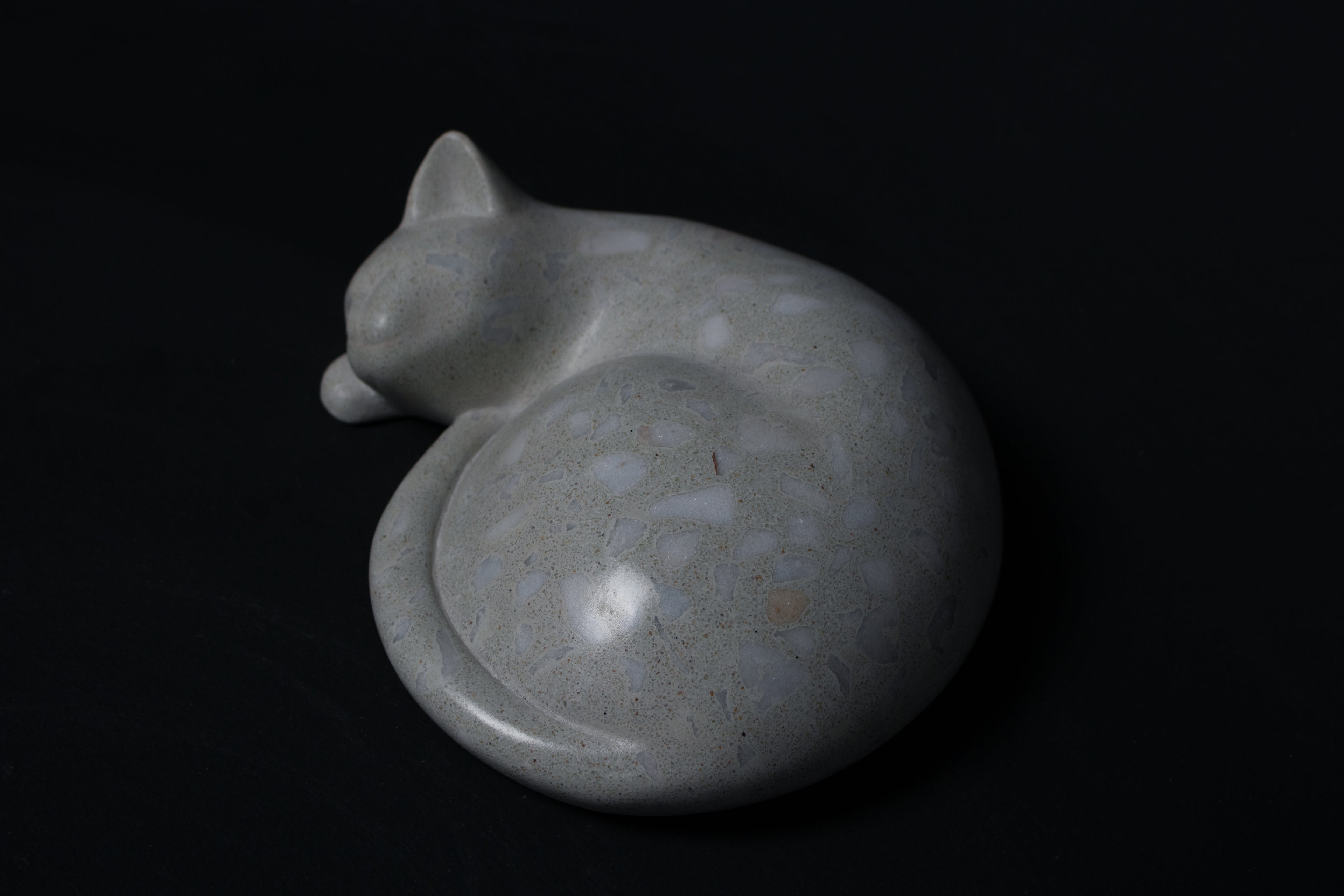 The Curly Cat terrazzo doorstop. A close-up view reveals the smoothly cast sleeping cat form, head resting on tucked paws, eyes closed, body curled into a low rounded mass. The pale grey terrazzo surface is flecked with irregular fragments of white and blush quartz.