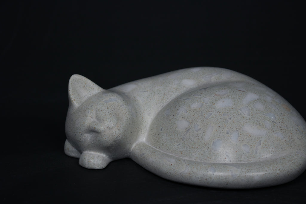 The Curly Cat terrazzo doorstop. A close-up view reveals the smoothly cast sleeping cat form, head resting on tucked paws, eyes closed, body curled into a low rounded mass. The pale grey terrazzo surface is flecked with irregular fragments of white and blush quartz.