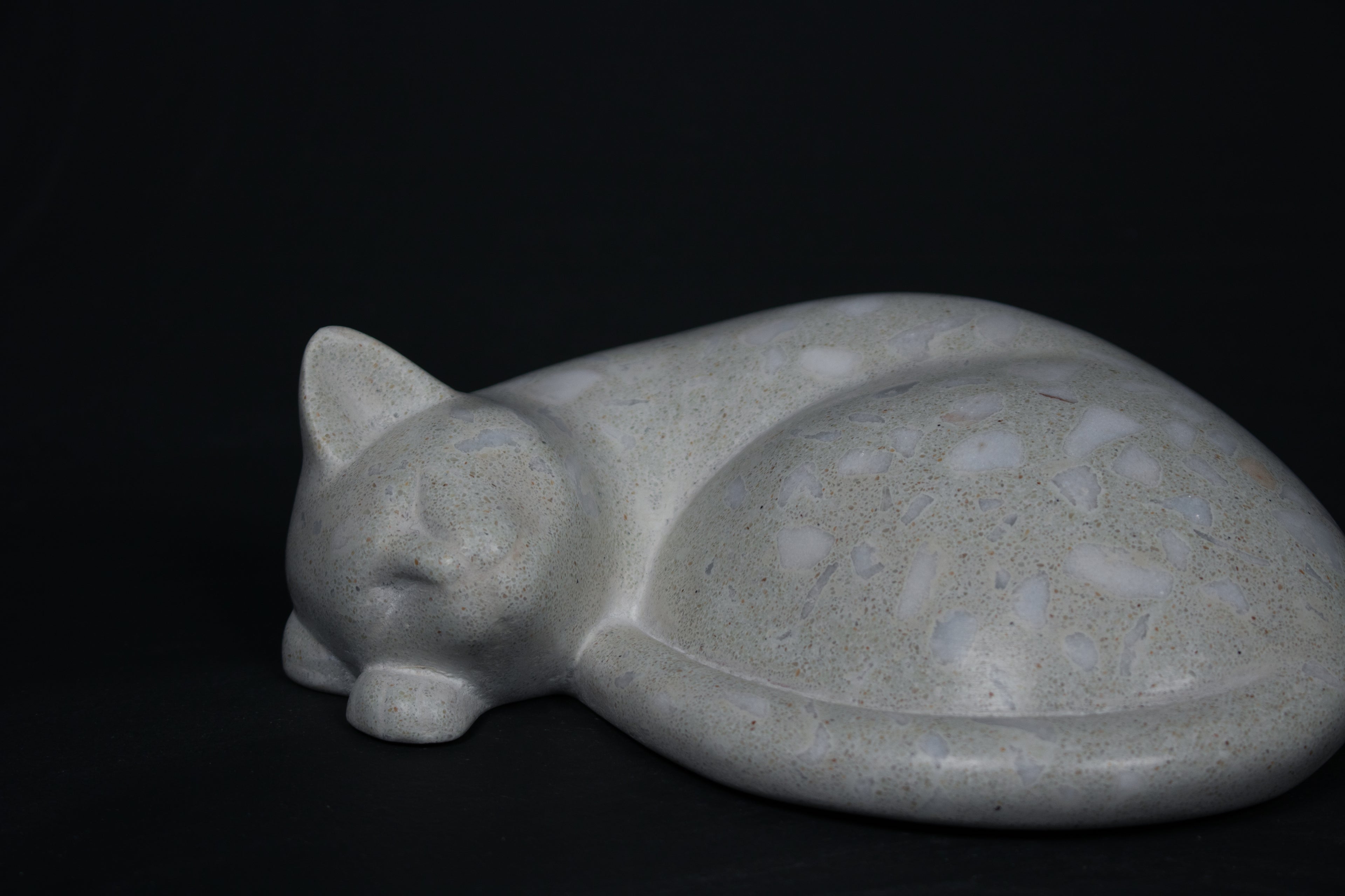The Curly Cat terrazzo doorstop. A close-up view reveals the smoothly cast sleeping cat form, head resting on tucked paws, eyes closed, body curled into a low rounded mass. The pale grey terrazzo surface is flecked with irregular fragments of white and blush quartz.