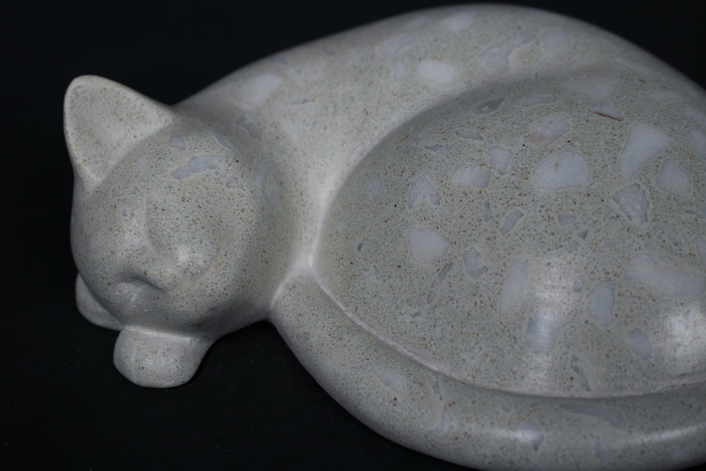 The Curly Cat terrazzo doorstop. A close-up view reveals the smoothly cast sleeping cat form, head resting on tucked paws, eyes closed, body curled into a low rounded mass. The pale grey terrazzo surface is flecked with irregular fragments of white and blush quartz.