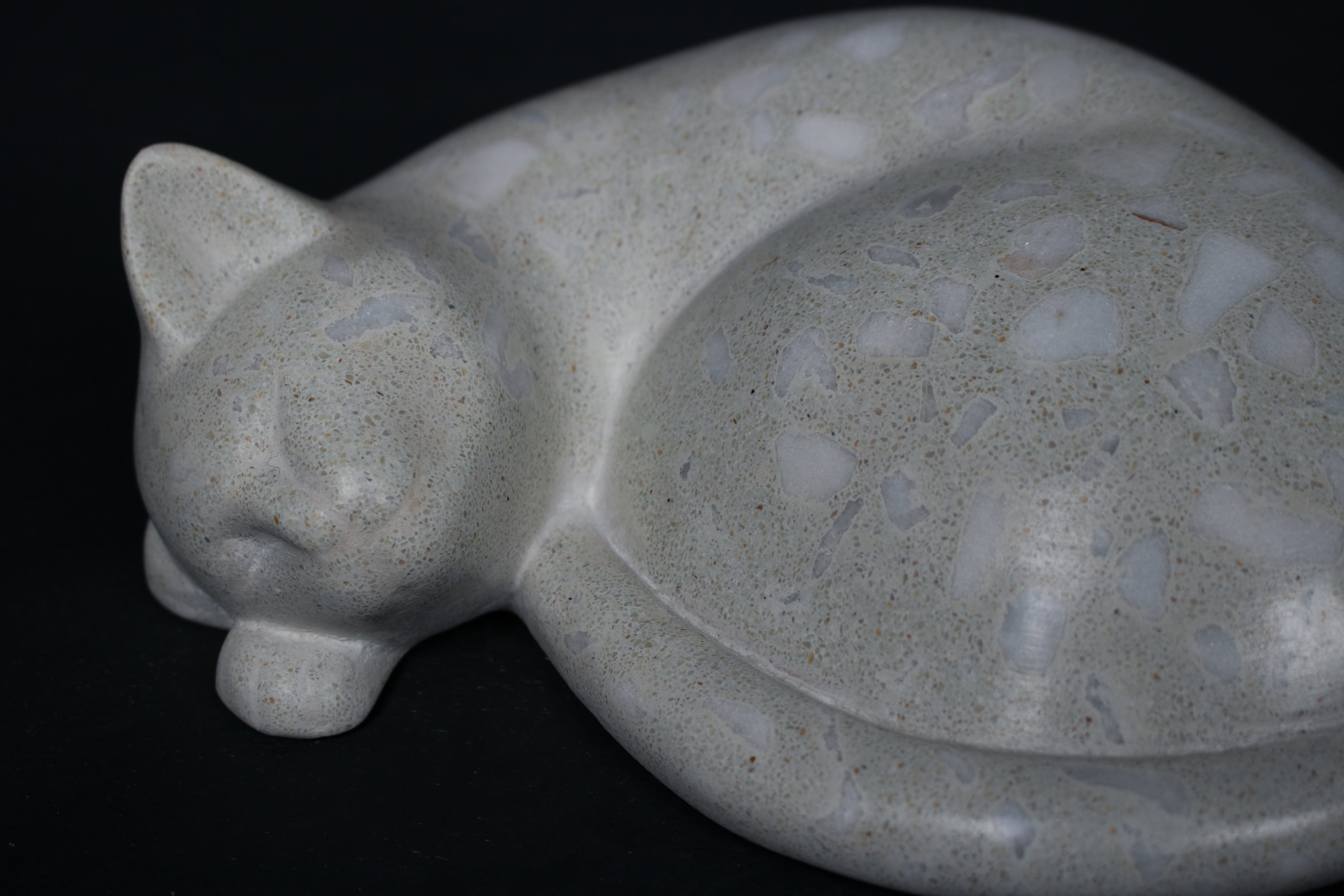 The Curly Cat terrazzo doorstop. A close-up view reveals the smoothly cast sleeping cat form, head resting on tucked paws, eyes closed, body curled into a low rounded mass. The pale grey terrazzo surface is flecked with irregular fragments of white and blush quartz.