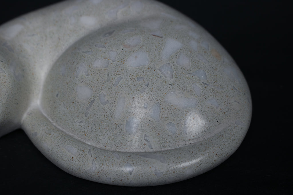The Curly Cat terrazzo doorstop. A close-up view reveals the smoothly cast sleeping cat form, head resting on tucked paws, eyes closed, body curled into a low rounded mass. The pale grey terrazzo surface is flecked with irregular fragments of white and blush quartz.