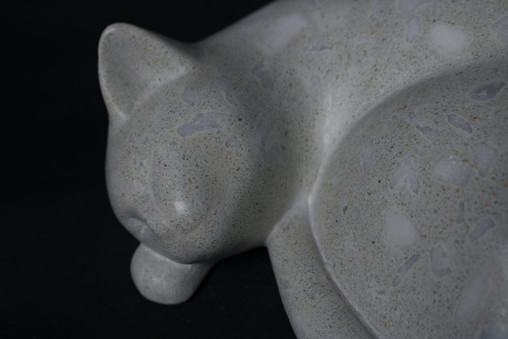 The Curly Cat terrazzo doorstop. A close-up view reveals the smoothly cast sleeping cat form, head resting on tucked paws, eyes closed, body curled into a low rounded mass. The pale grey terrazzo surface is flecked with irregular fragments of white and blush quartz.