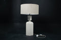 The Dacca table lamp. The cast stone base has a tall rectangular bottle form with chamfered edges, narrowing to a smaller square collar. A fabric-wrapped cord trails to a white inline switch.