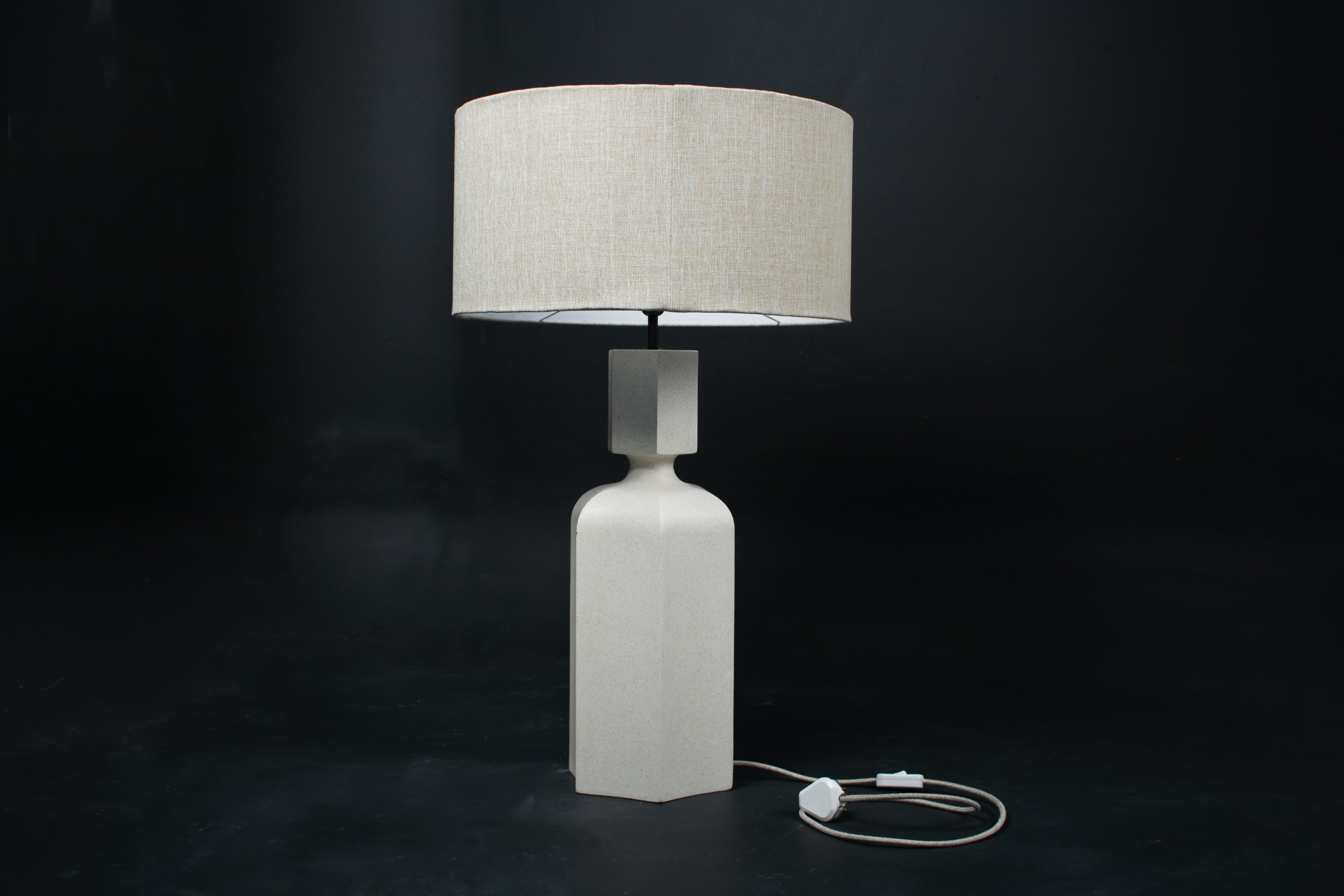 The Dacca table lamp. The cast stone base has a tall rectangular bottle form with chamfered edges, narrowing to a smaller square collar. A fabric-wrapped cord trails to a white inline switch.