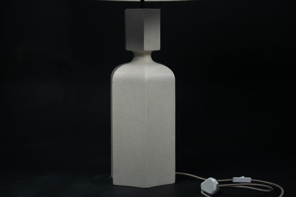 The Dacca table lamp. The cast stone base has a tall rectangular bottle form with chamfered edges, narrowing to a smaller square collar. A fabric-wrapped cord trails to a white inline switch.