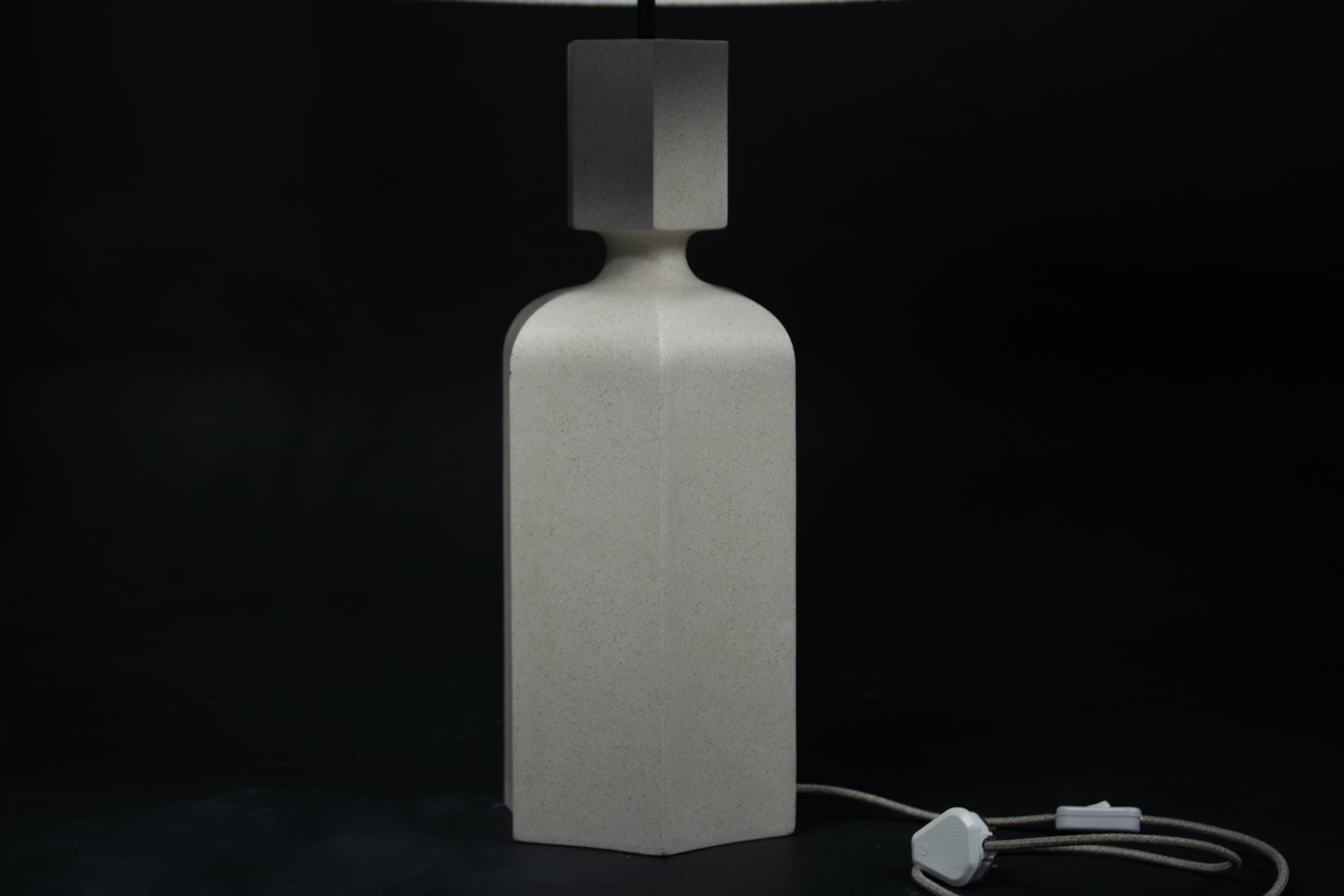 The Dacca table lamp. The cast stone base has a tall rectangular bottle form with chamfered edges, narrowing to a smaller square collar. A fabric-wrapped cord trails to a white inline switch.