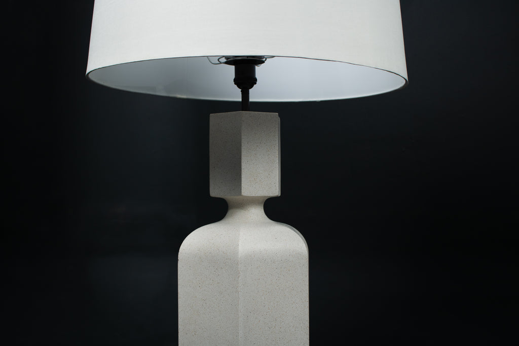 The Dacca table lamp. The cast stone base has a tall rectangular bottle form with chamfered edges, narrowing to a smaller square collar. A fabric-wrapped cord trails to a white inline switch.