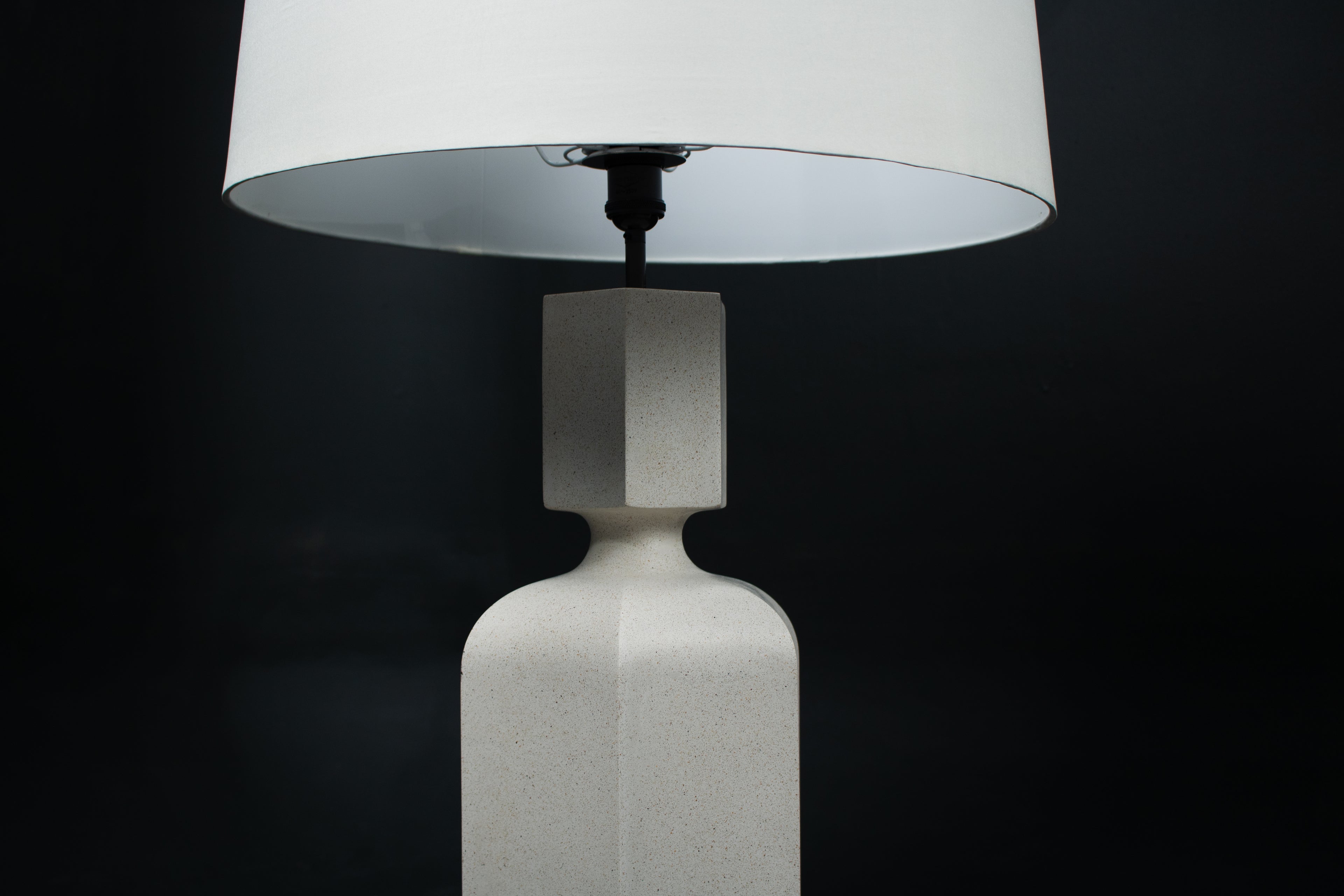 The Dacca table lamp. The cast stone base has a tall rectangular bottle form with chamfered edges, narrowing to a smaller square collar. A fabric-wrapped cord trails to a white inline switch.