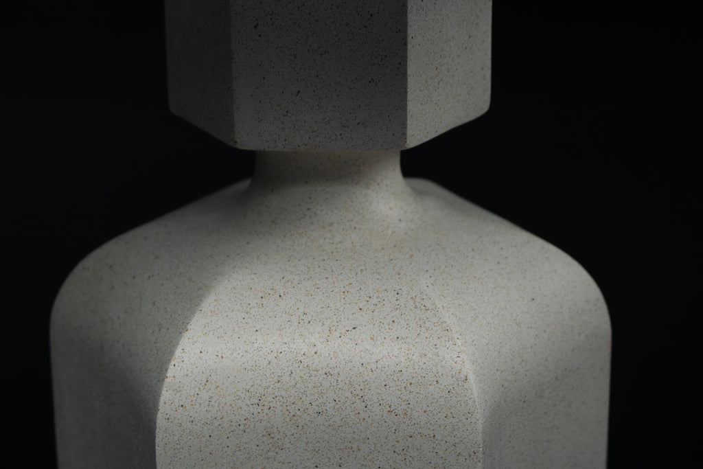 The Dacca table lamp. The cast stone base has a tall rectangular bottle form with chamfered edges, narrowing to a smaller square collar. A fabric-wrapped cord trails to a white inline switch.