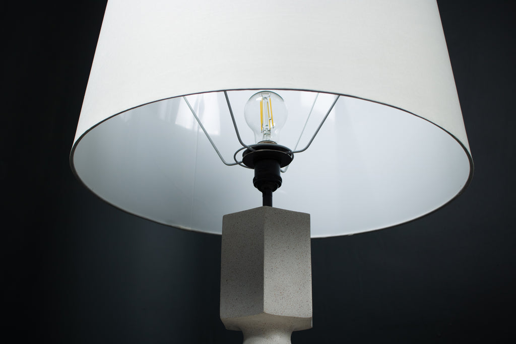 The Dacca table lamp. The cast stone base has a tall rectangular bottle form with chamfered edges, narrowing to a smaller square collar. A fabric-wrapped cord trails to a white inline switch.