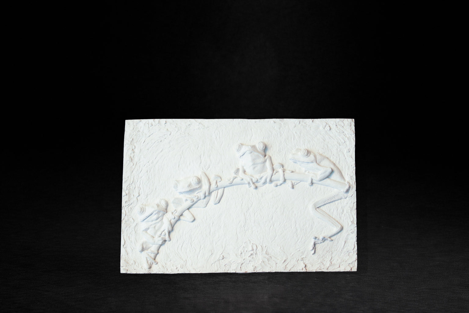 White plaster relief panel depicting four red-eyed tree frogs clinging to a branch.