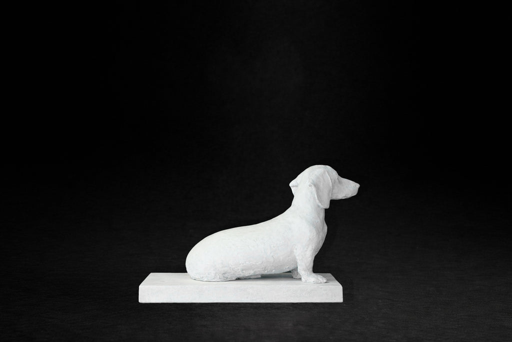 White plaster sculpture of a seated dachshund on a rectangular base, gazing forward.