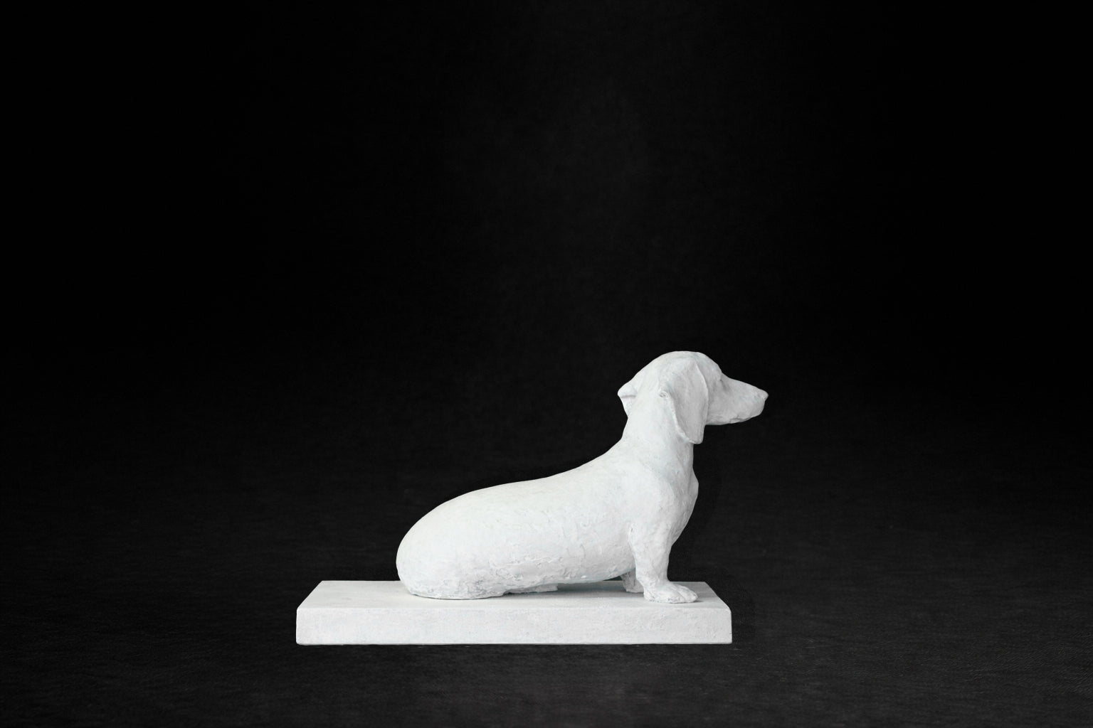 White plaster sculpture of a seated dachshund on a rectangular base, gazing forward.