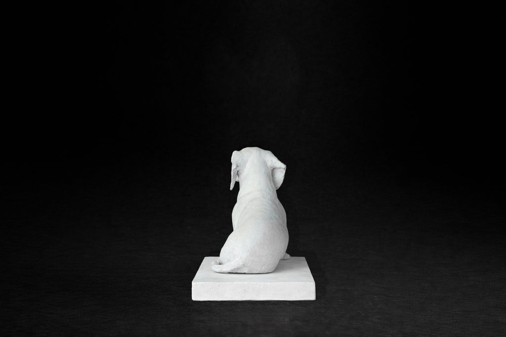 White plaster sculpture of a seated dachshund on a rectangular base, gazing forward.