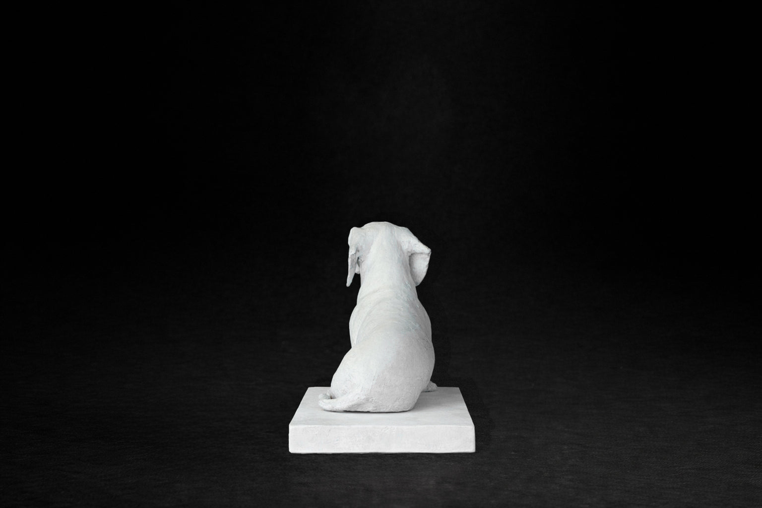 White plaster sculpture of a seated dachshund on a rectangular base, gazing forward.