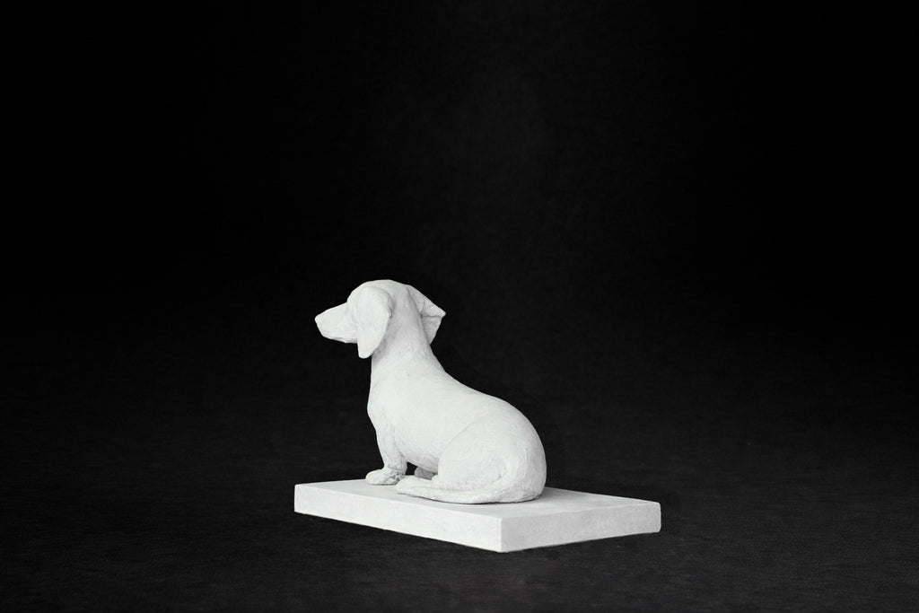White plaster sculpture of a seated dachshund on a rectangular base, gazing forward.