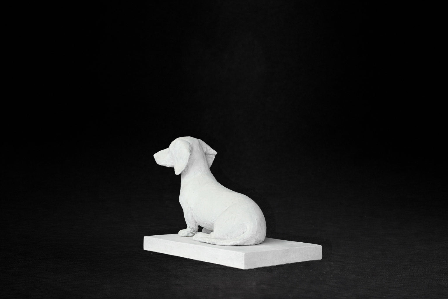 White plaster sculpture of a seated dachshund on a rectangular base, gazing forward.