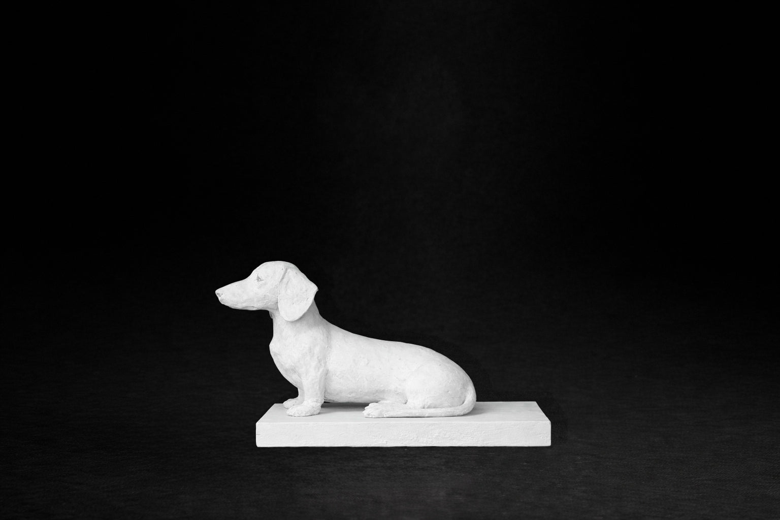 White plaster sculpture of a seated dachshund on a rectangular base, gazing forward.