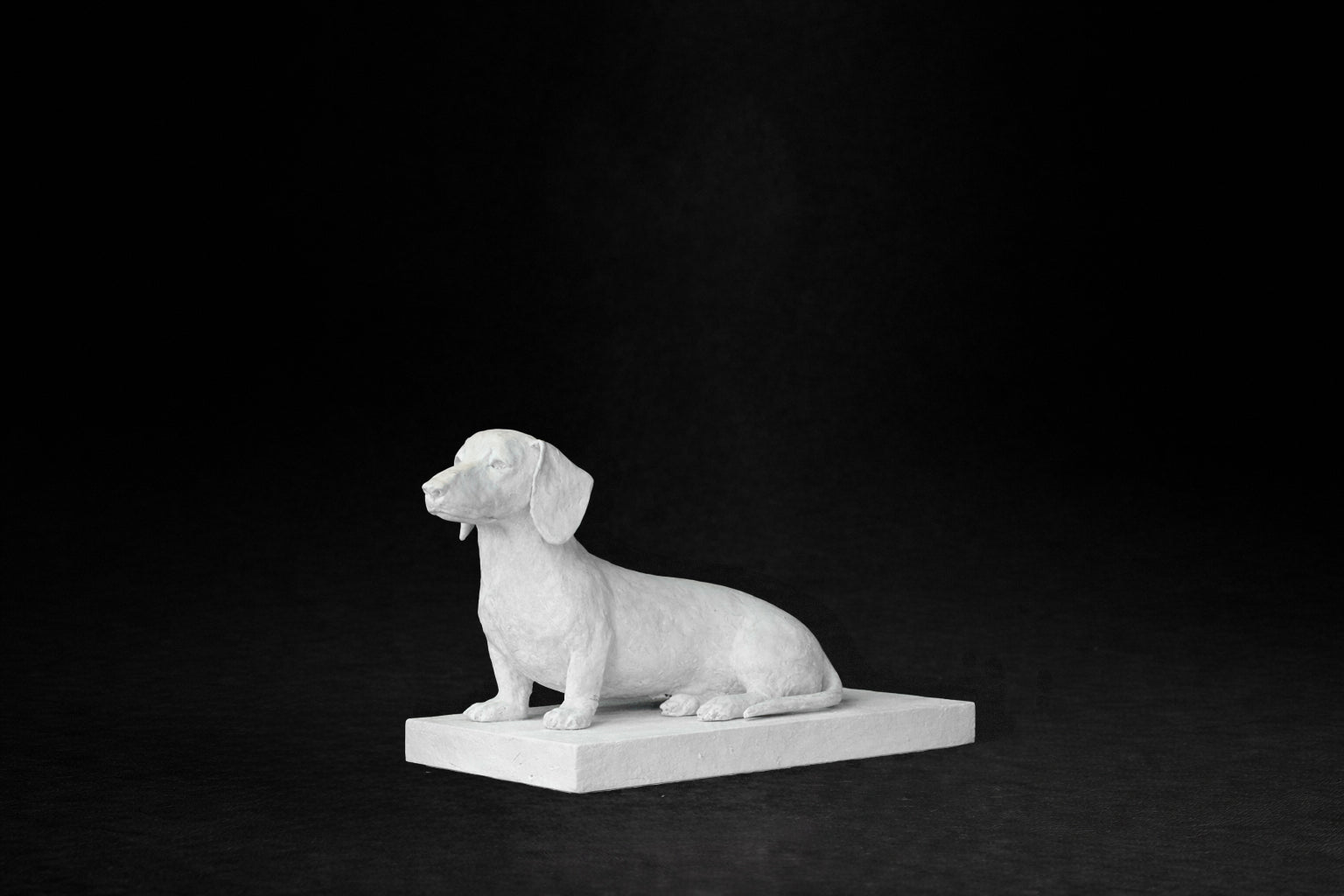 White plaster sculpture of a seated dachshund on a rectangular base, gazing forward.