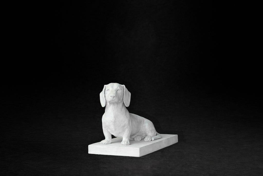 White plaster sculpture of a seated dachshund on a rectangular base, gazing forward.