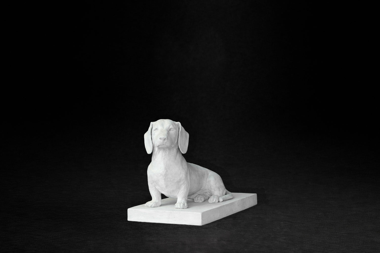 White plaster sculpture of a seated dachshund on a rectangular base, gazing forward.